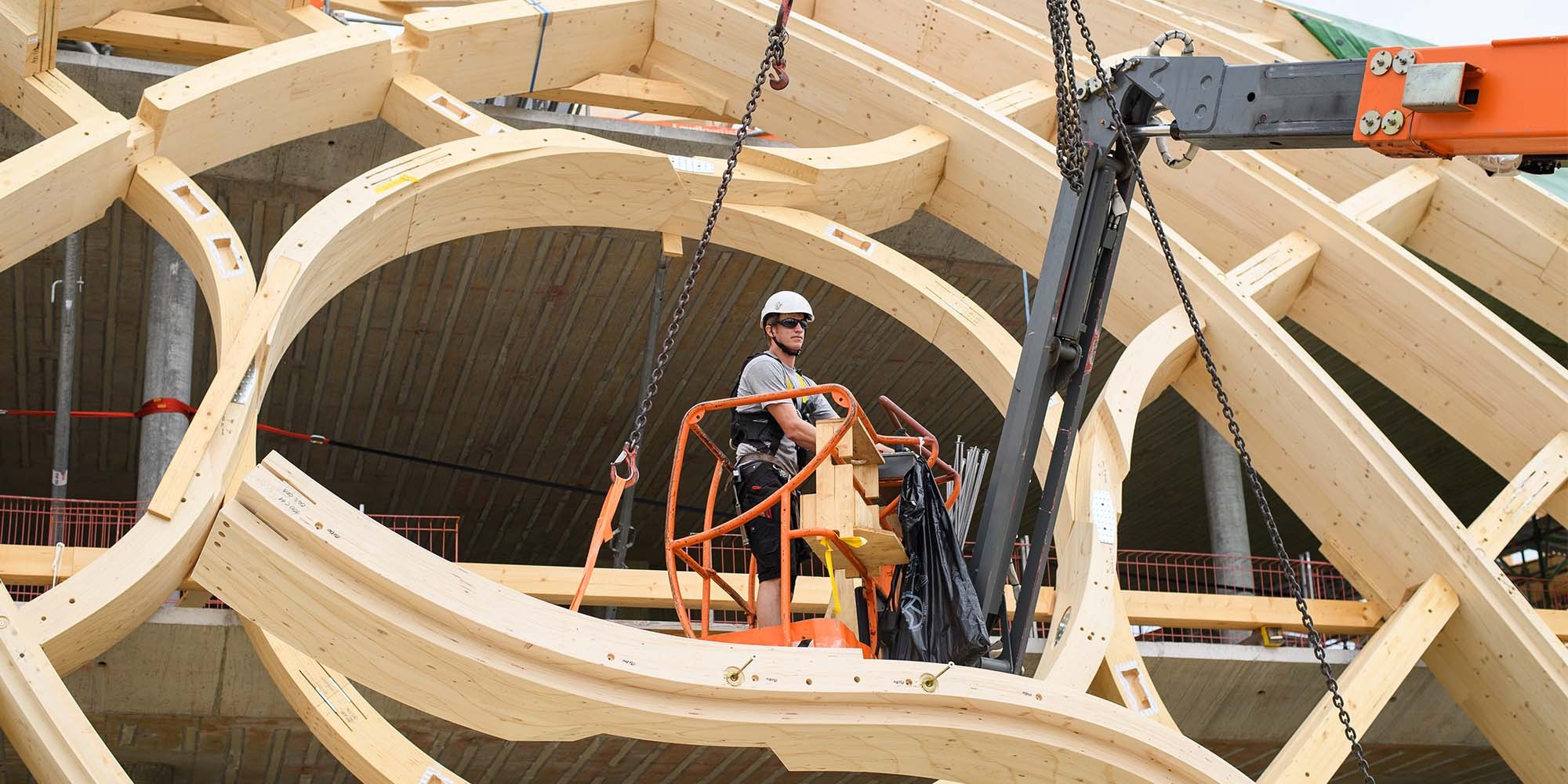 Ein Montagearbeiter befindet sich mit einem orangen Hebekran inmitten der Holzkonstruktion des Swatch-Gebäudes