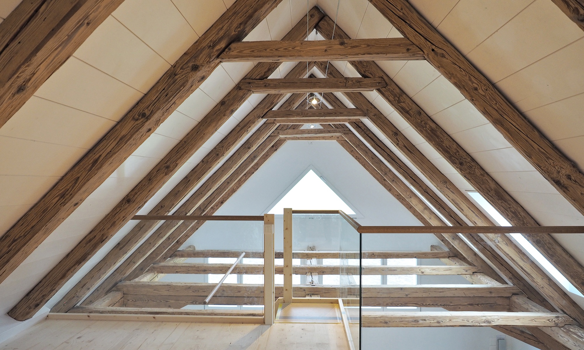 Lichtdurchflutetes offenes Dachgeschoss gesichert durch Glas- und Holzgeländer. Die Dachschrägen sind verkleidet mit breiten Holzbalken.