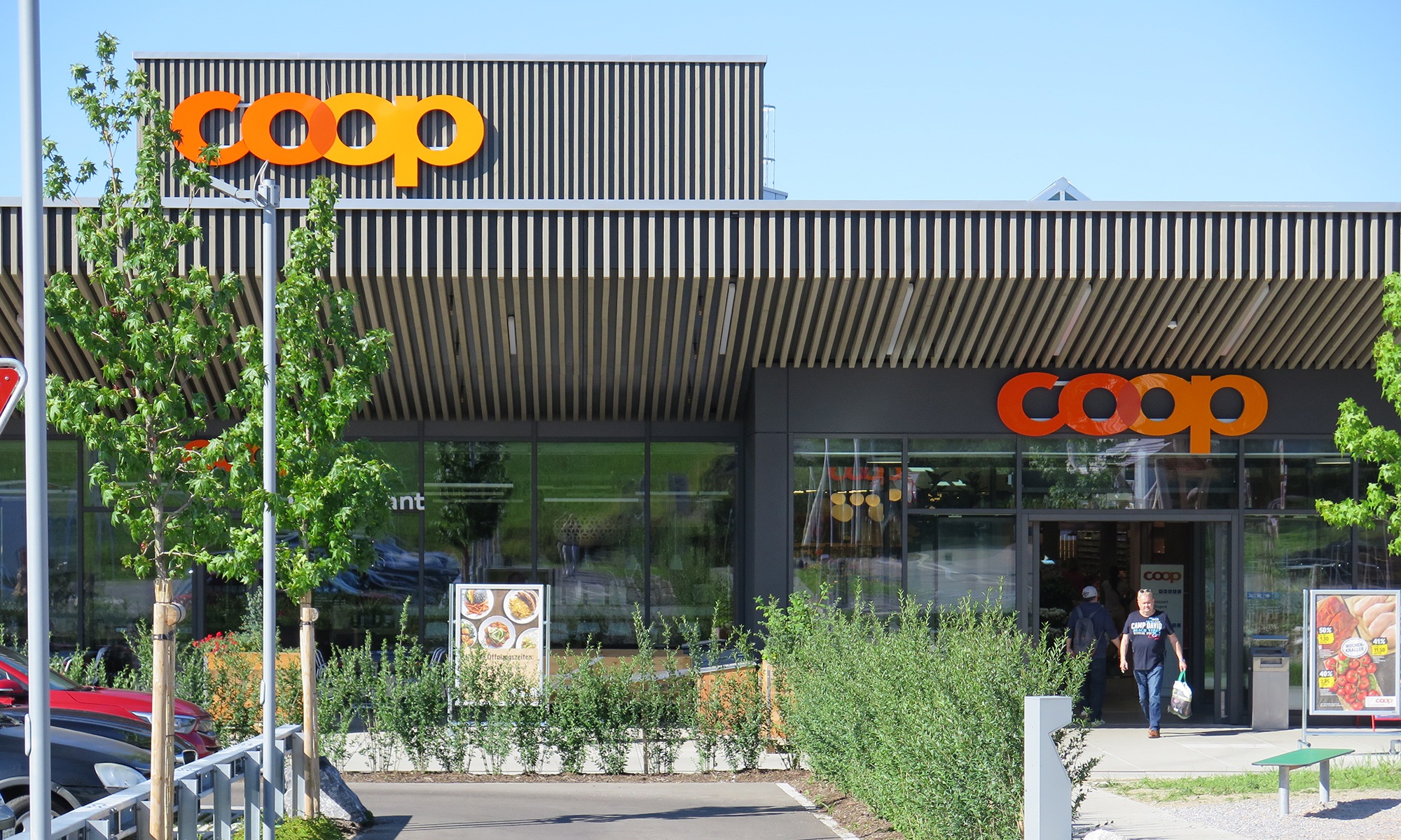 Haupteingang des Coop Super Center in Uzwil mit dunkler Holzfassade sowie Vorplatz, kleinen Bäumen und Kleinpflanzen.