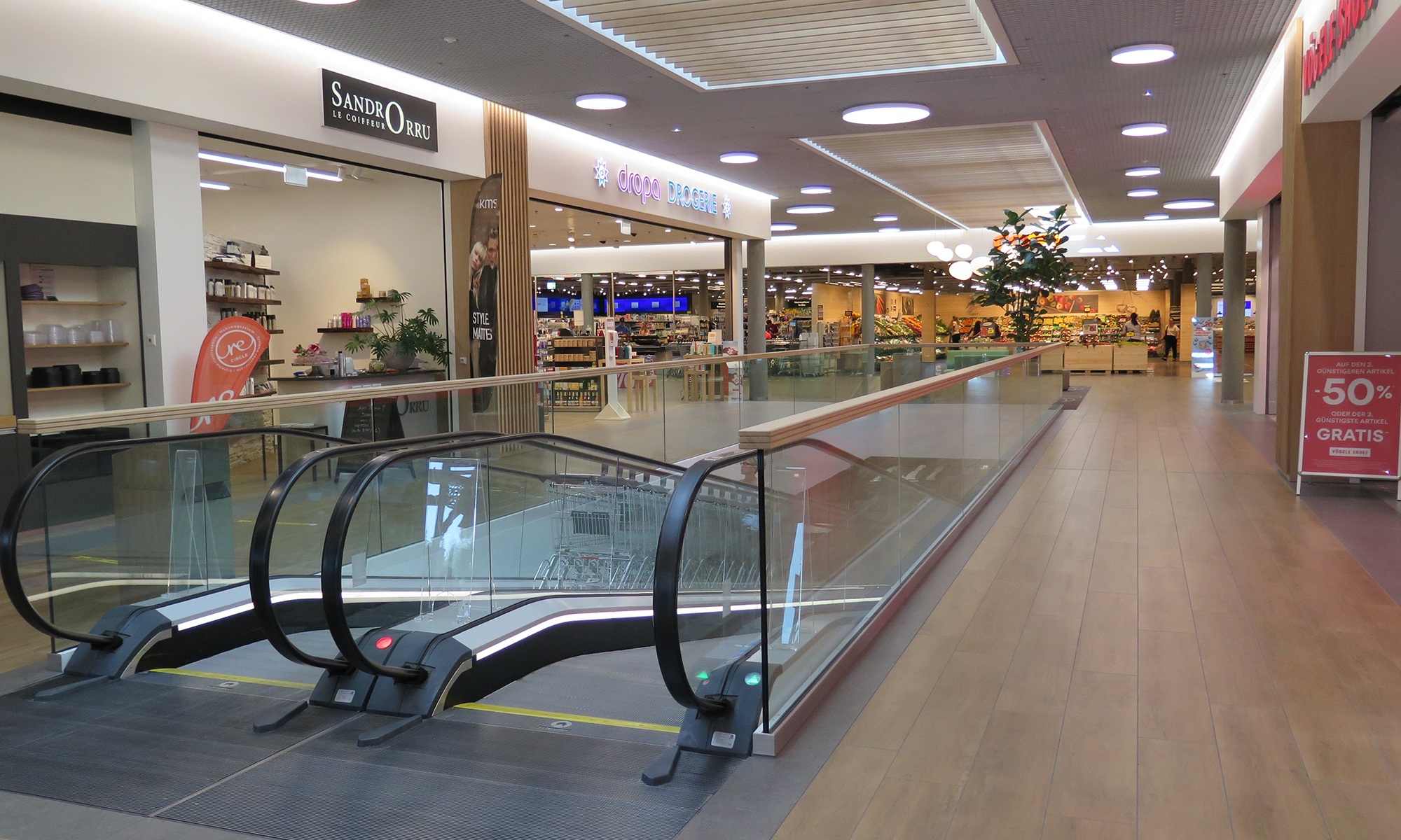 Im Eingangsbereich des Coop Super Centers Uzwil mit den Eingängen zu den Geschäften sowie Rolltreppen und Fussboden aus hellem Holz.