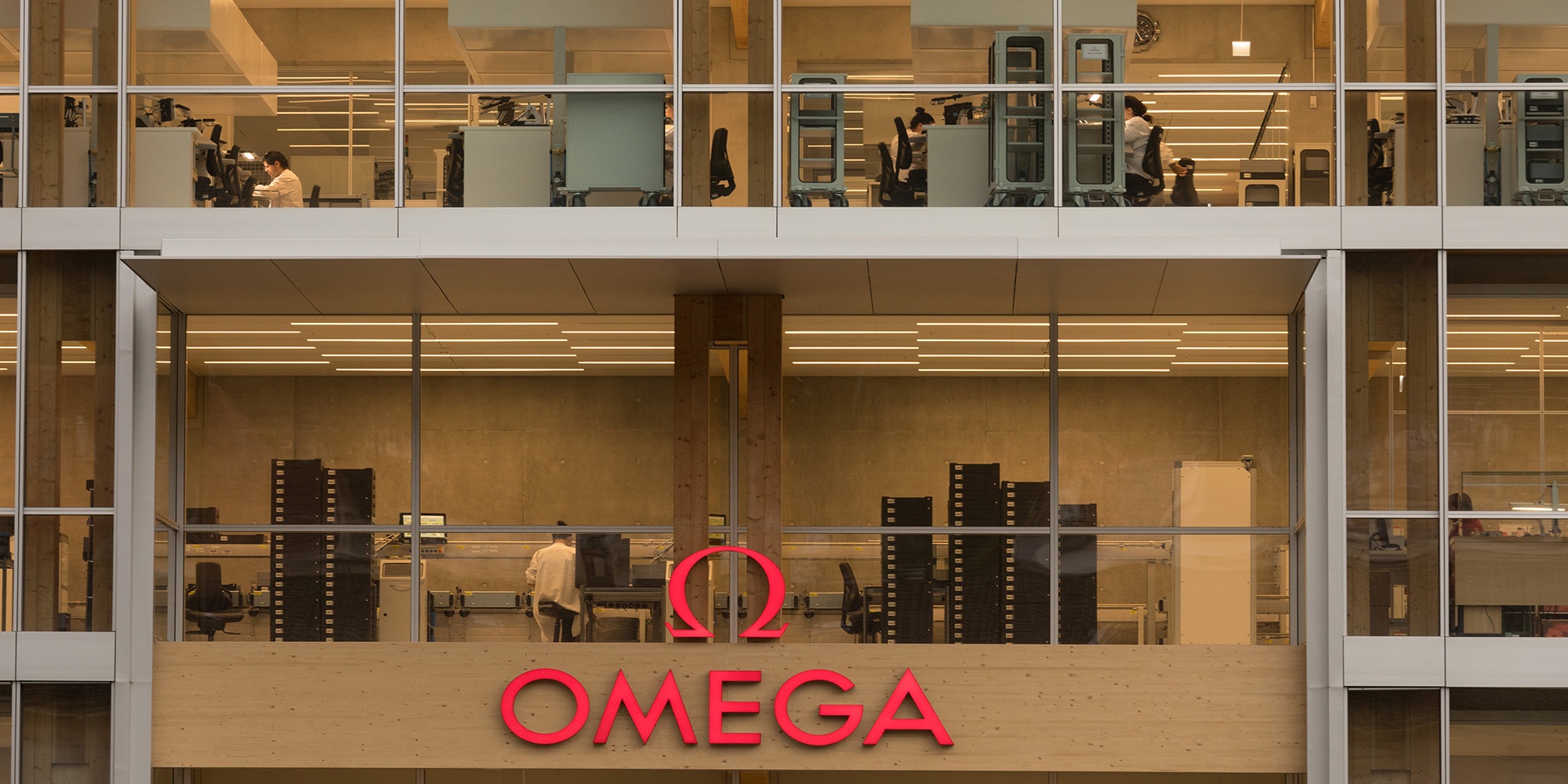 Grosse Omega Schrift in Rot am neuen Bürogebäude aus Holz mit Glasfront. Im Hintergrund sieht man Mitarbeitende an ihren Arbeitsplätzen. 