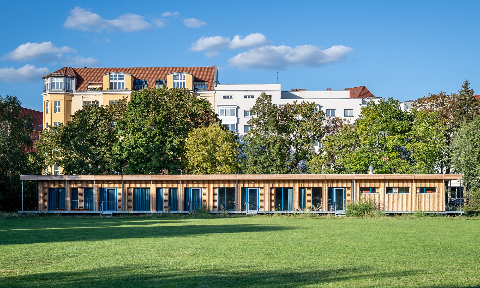 Visualisierung eines eingeschossiges temporären Schulhauses in Berlin Schönefeld. Die Holzfassade hebt sich von der grünen Wiese und vom blauen Himmel ab.
