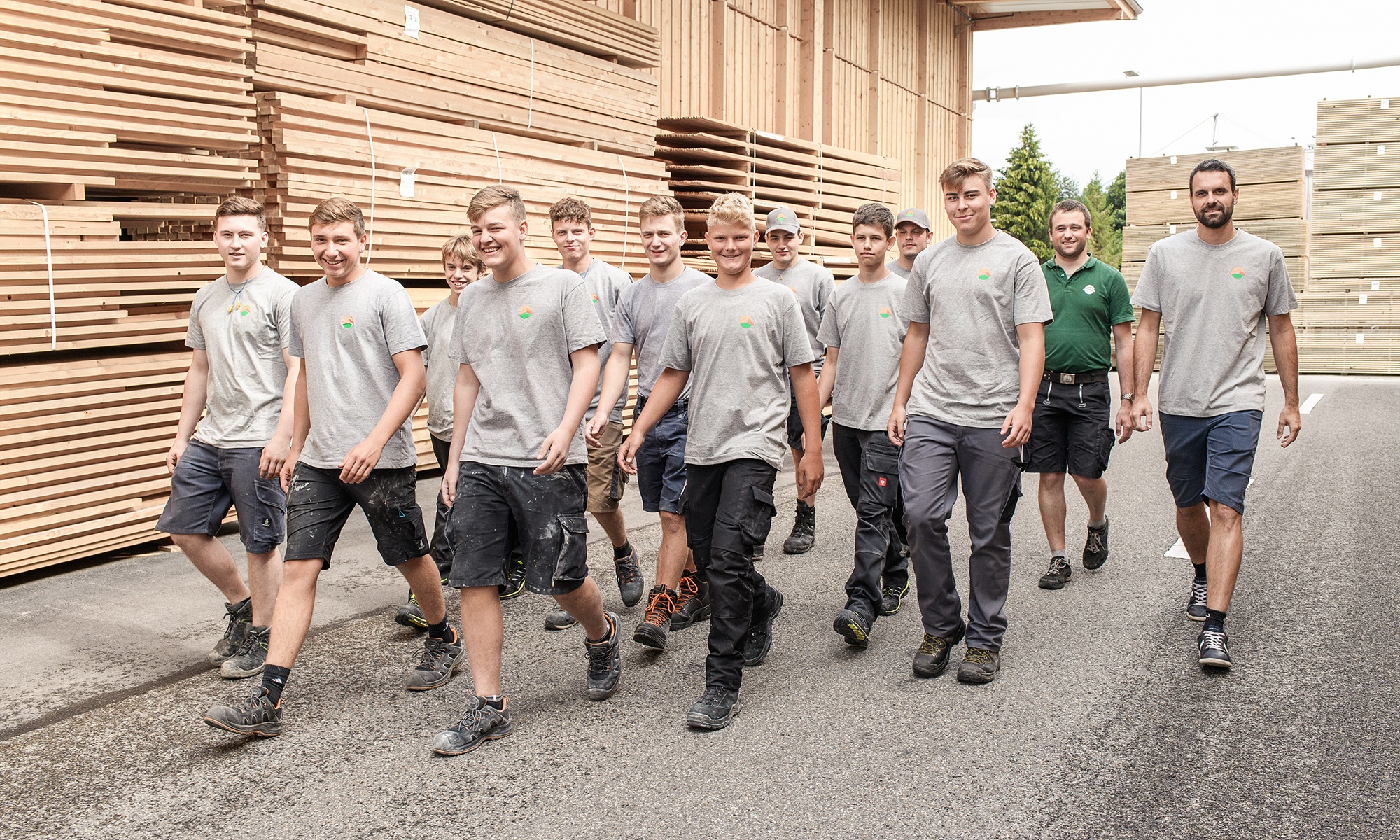 Ein Gruppenbild mit elf jungen Leuten in der Ausbildung zum Zimmermann und zwei Ausbildner laufen vor grossen Holzstapel durch und lachen