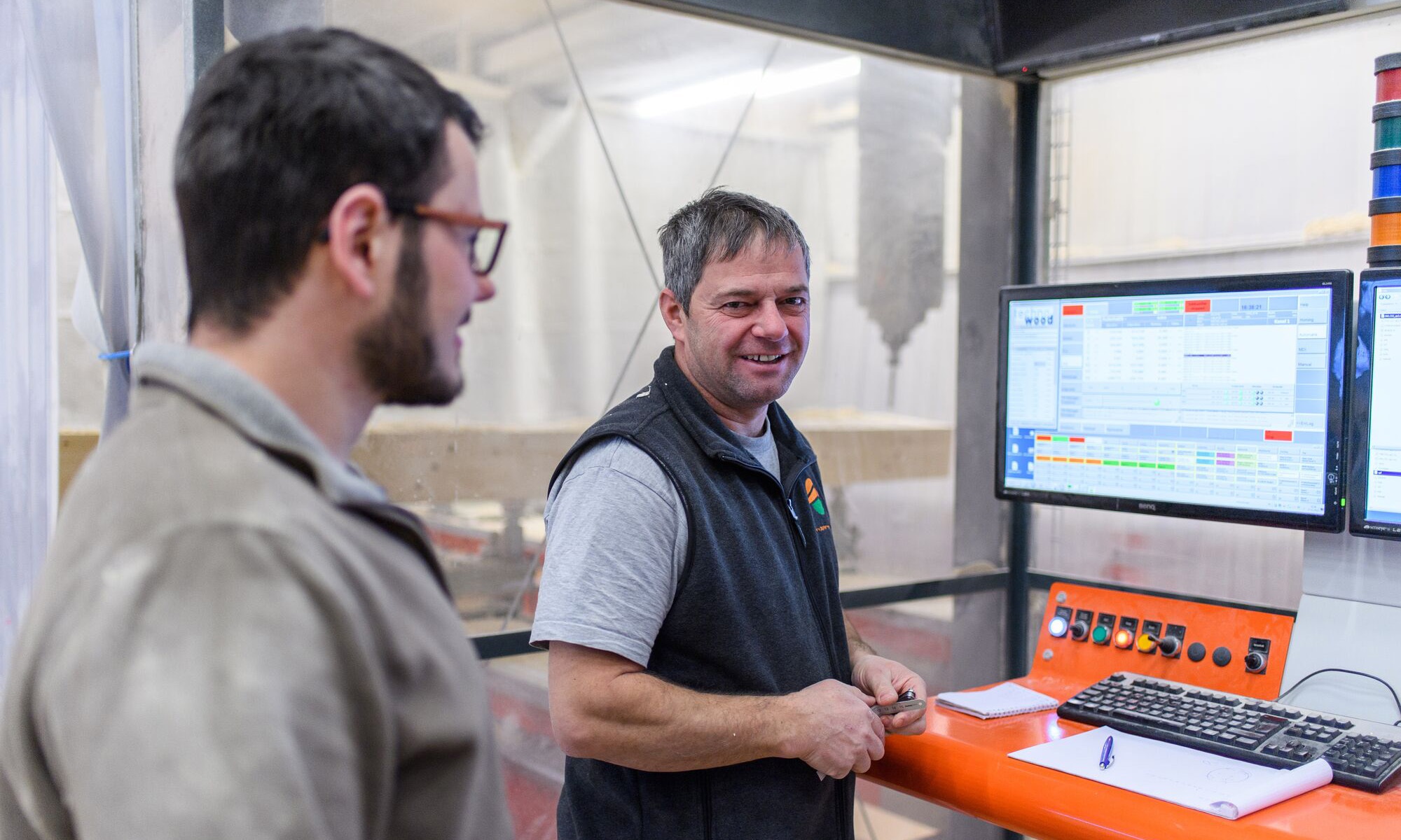 2 people at the controls of a CNC machine. 2 PCs are being used in the background. One person is looking at the camera, the other at the screen.