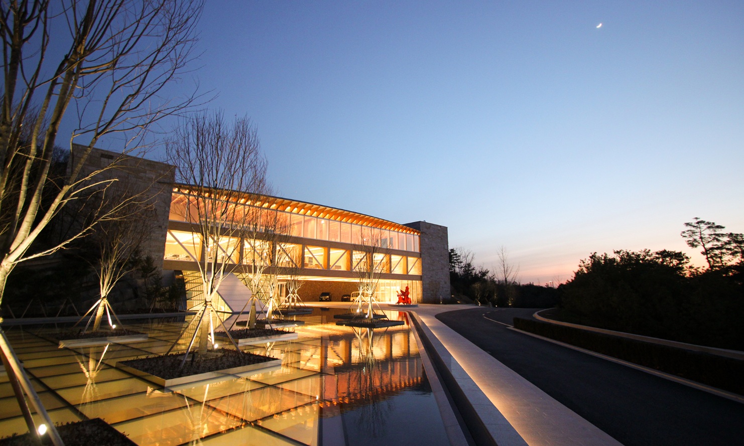 External photograph of the Haesley Nine Bridges Golf Resort Learning Centre. Illuminated timber architecture at night, with the pool in the foreground.<br/>