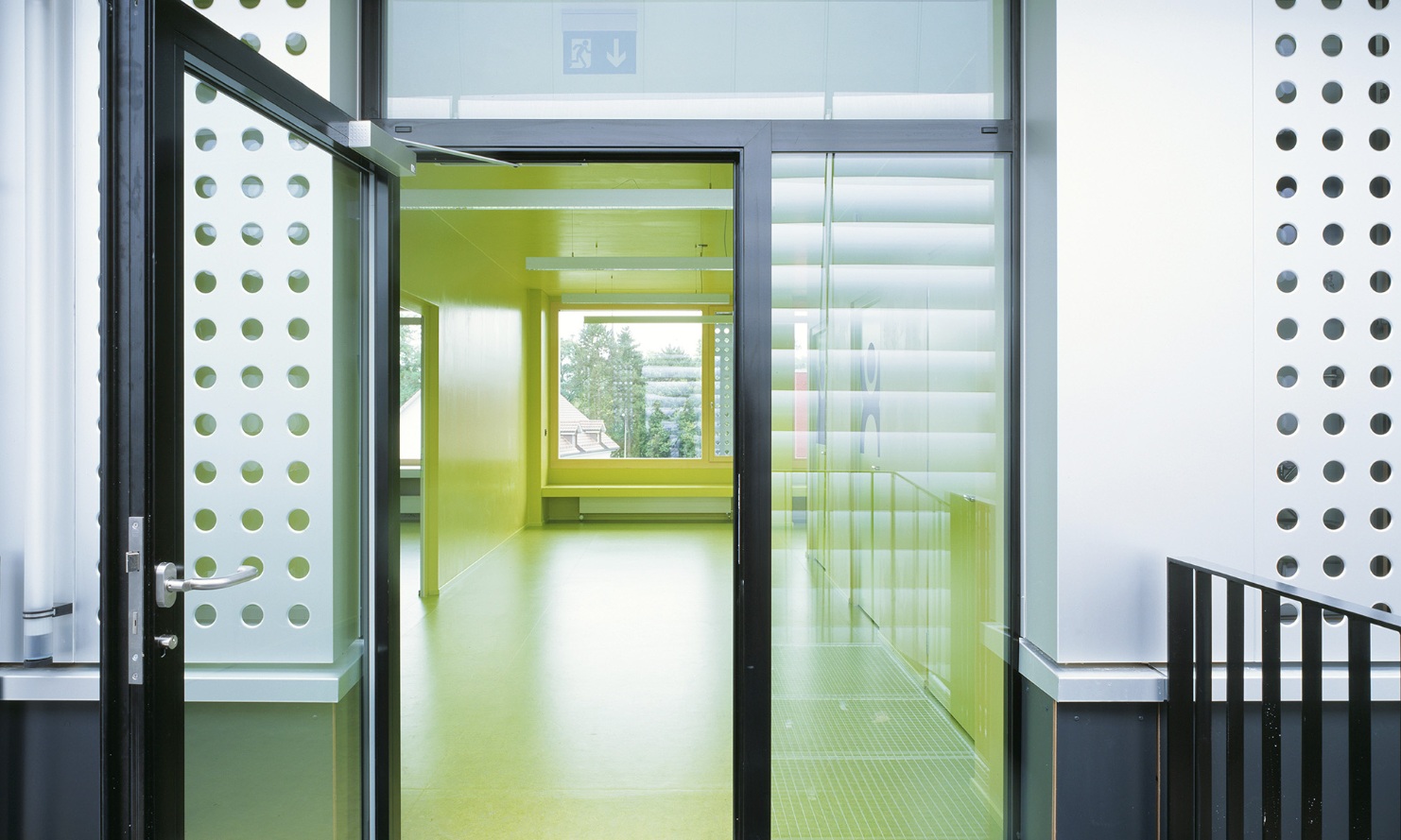 Espace d’entrée au design attrayant du pavillon provisoire de l’école à Männedorf avec porte vitrée et sol jaune dans la zone d’entrée.