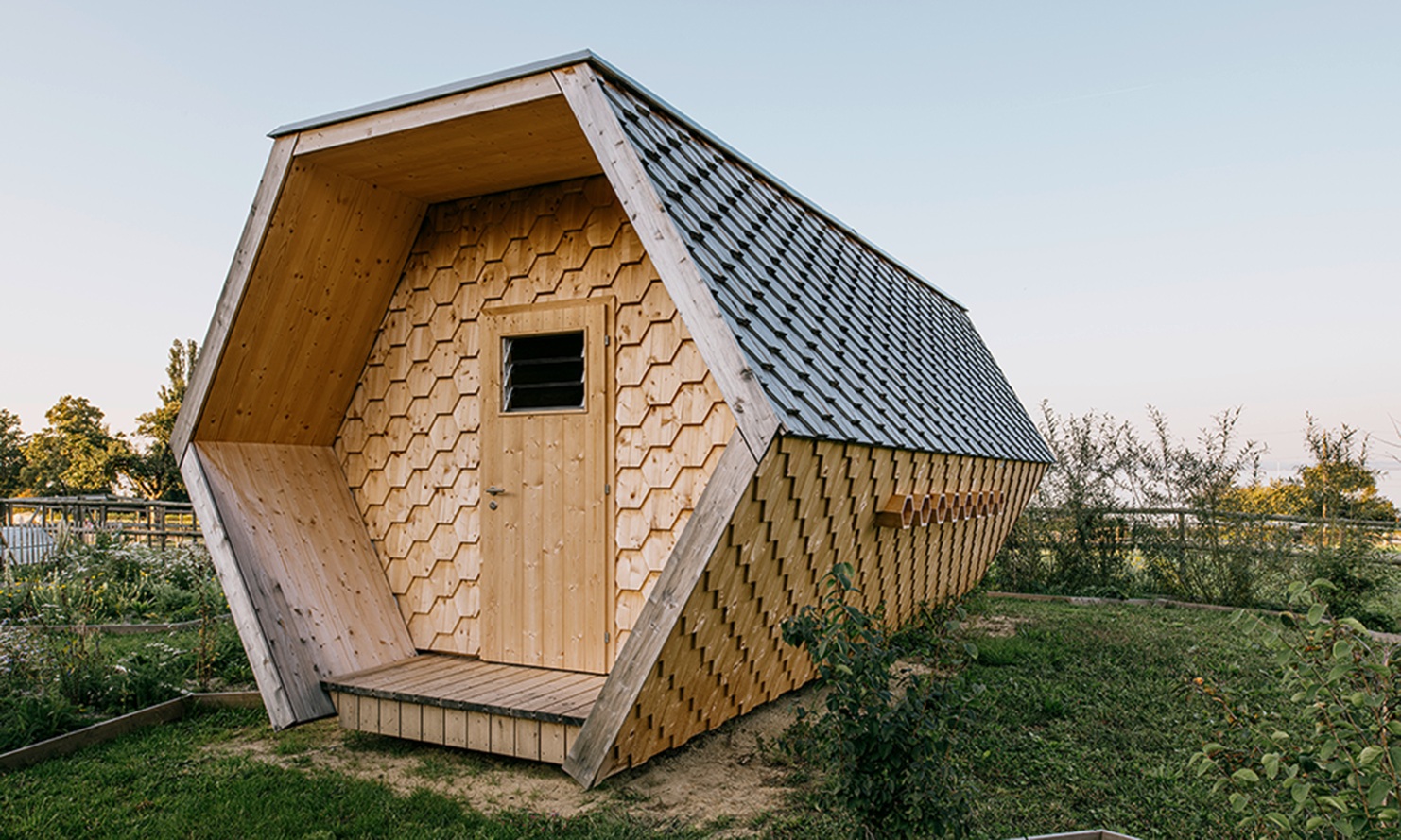 Honeycomb-shaped bee house with its facade of hexagonal wood shingles nestled in a flowering meadow.