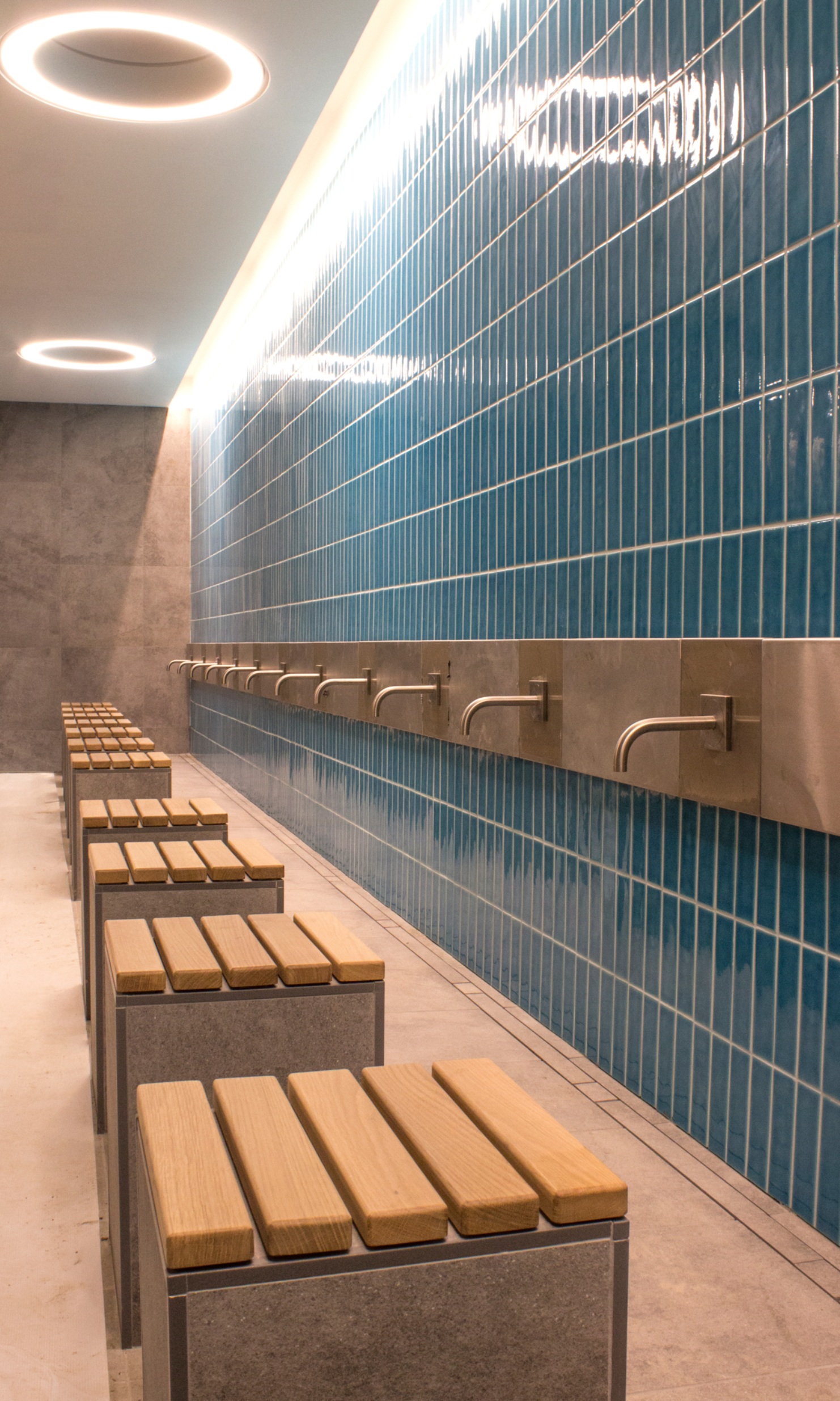 The photograph shows the washing area of the Cambridge Mosque. The stools are made of stone and clad with wooden panels. The taps are fixed to a blue slab wall.