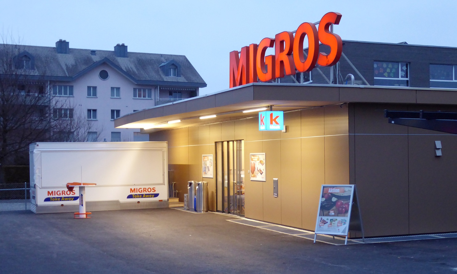 A single-storey temporary timber construction at dusk with the Migros neon sign on the roof.