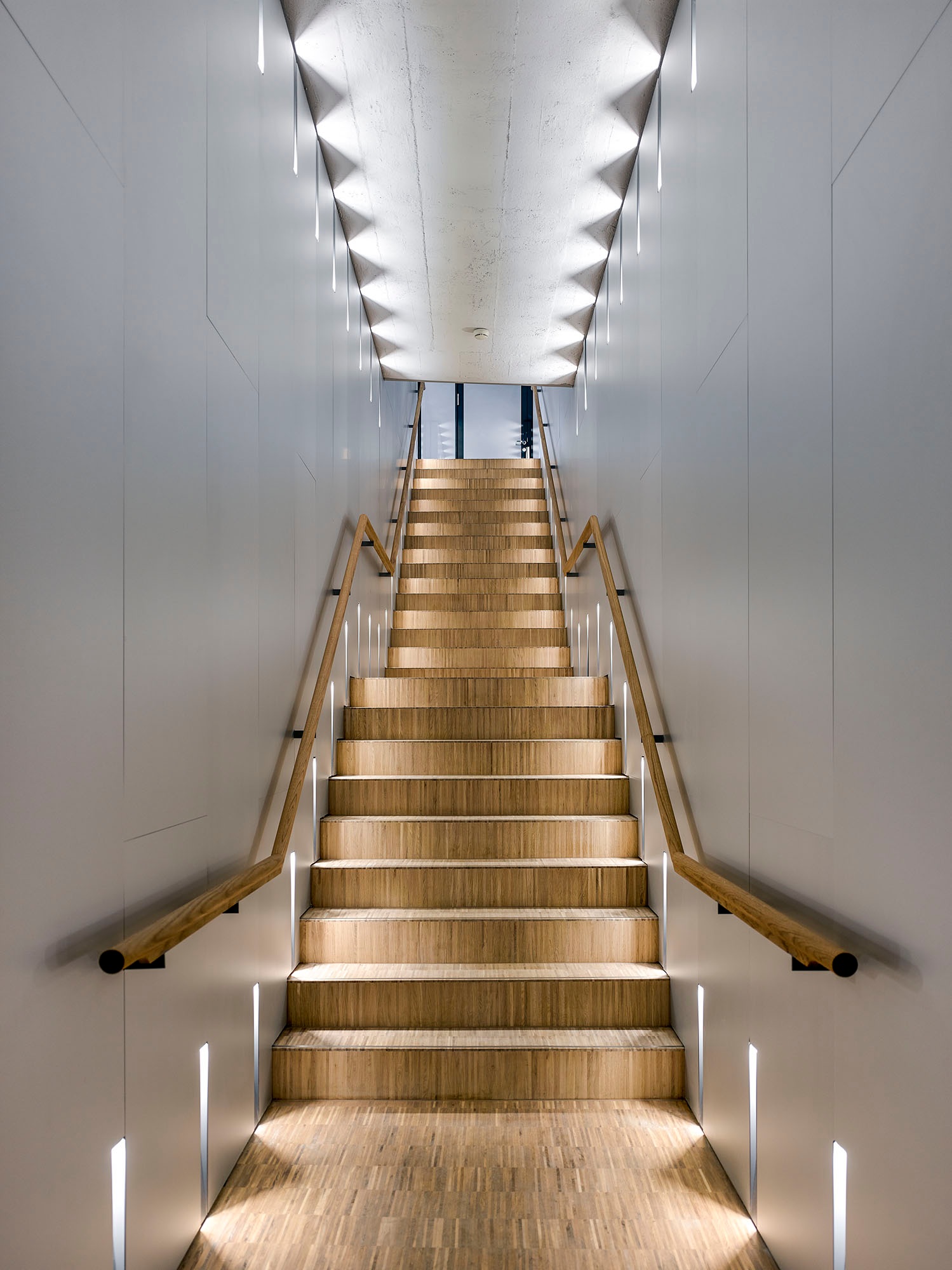 Un escalier recouvert de parquet mène à l’étage supérieur et se distingue de manière remarquable par rapport à la blancheur des murs et des plafonds.