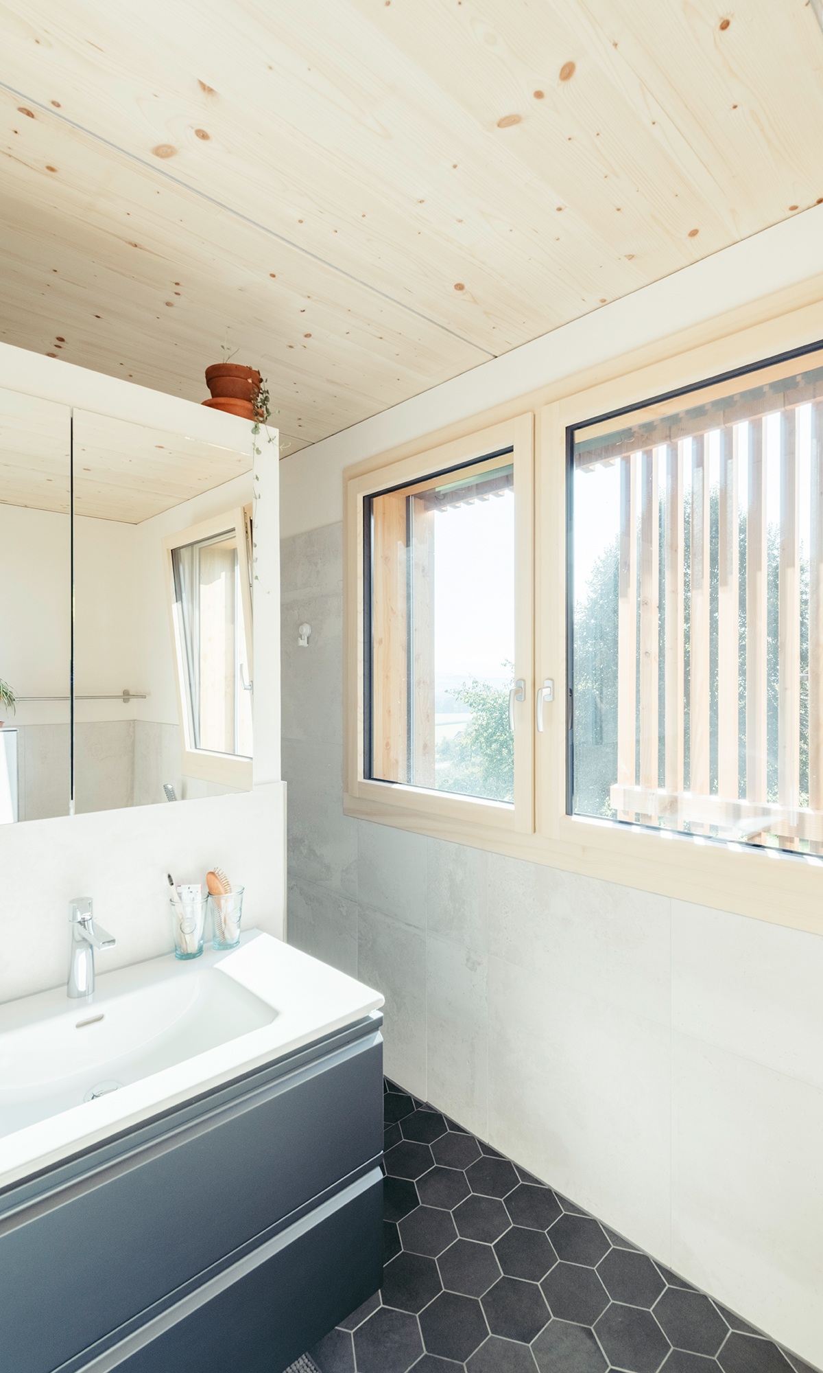 Viel natürliches Licht fällt durch die Fenster ins Badezimmer mit grauem Fussboden und Badmöbeln.