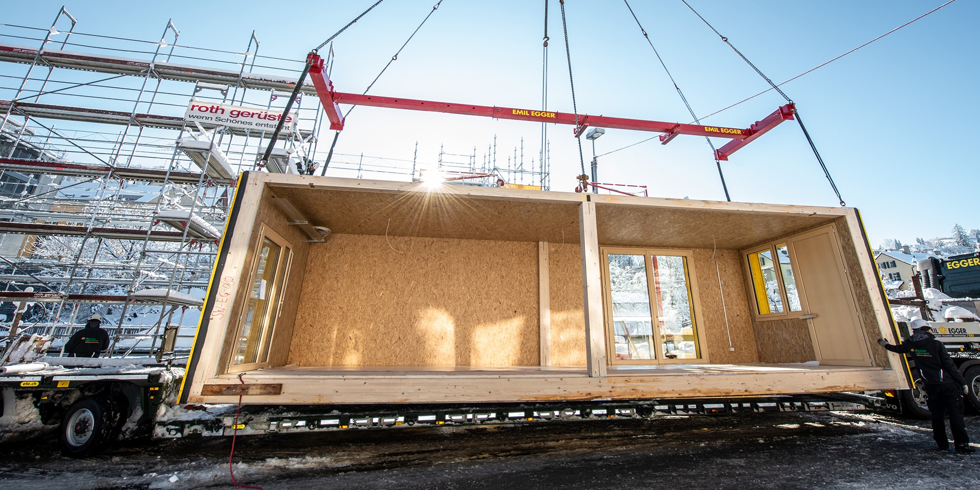 Timber module on a crane: truck is unloaded on the construction site in bright winter sunshine.