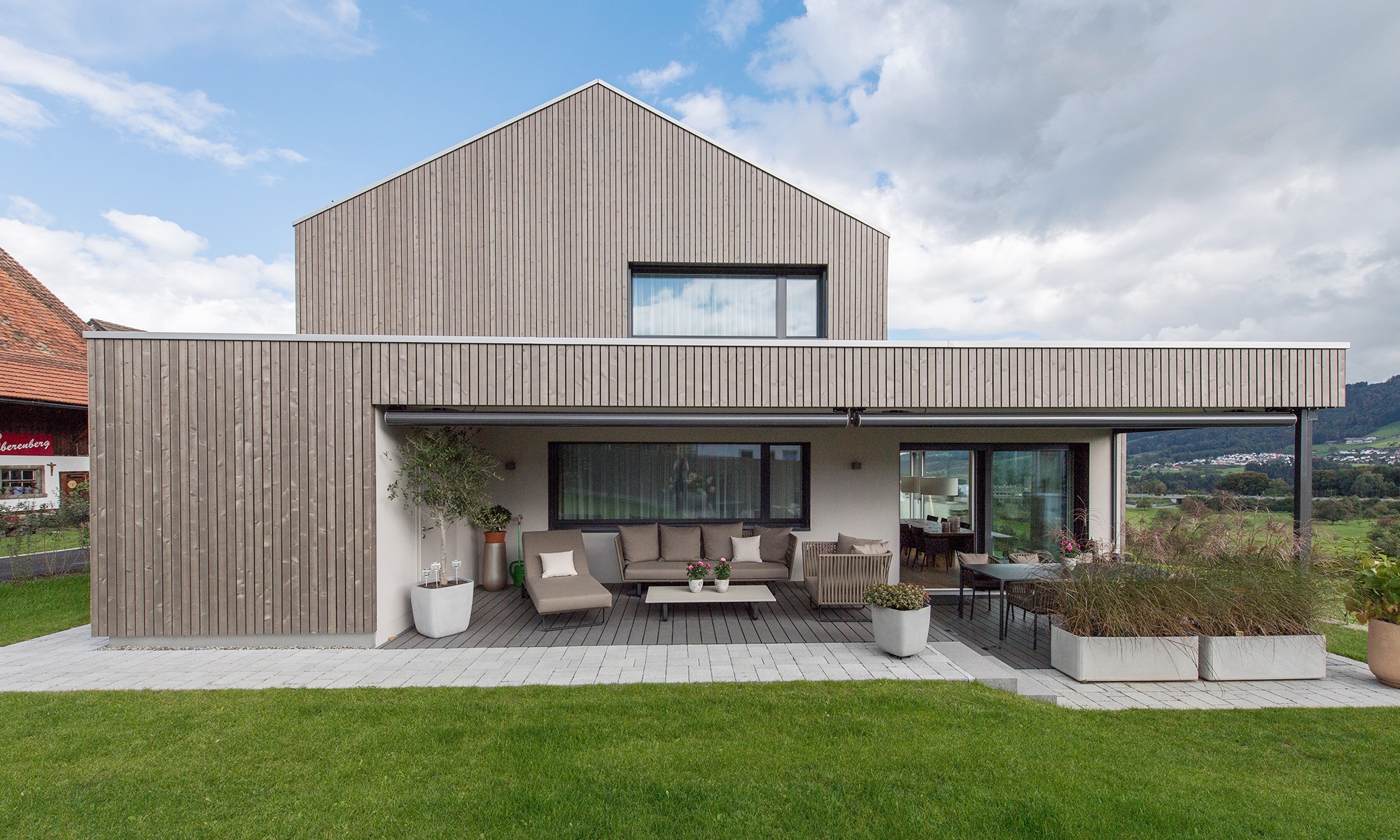 Aufnahme zeigt das Einfamilienhaus mit Seeblick sowie dessen überdachten Sitzplatz im Gartenbereich bei schönem Wetter