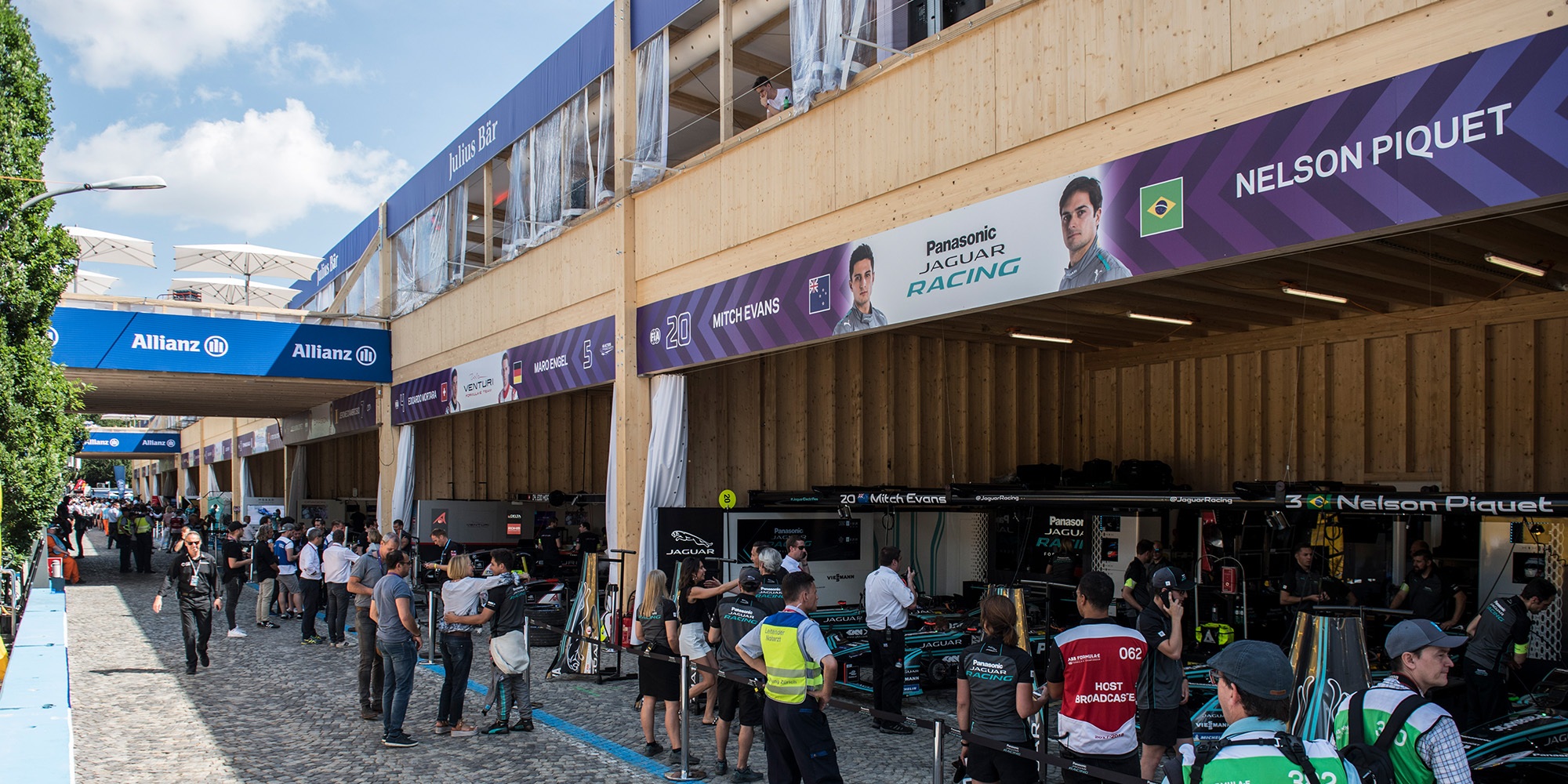 Le bâtiment événementiel en bois a servi de ligne des stands au Zurich E-Prix 2018. Au premier plan, on voit des visiteurs et des membres des équipes. En arrière-plan, les voitures de course sont dans les stands.
