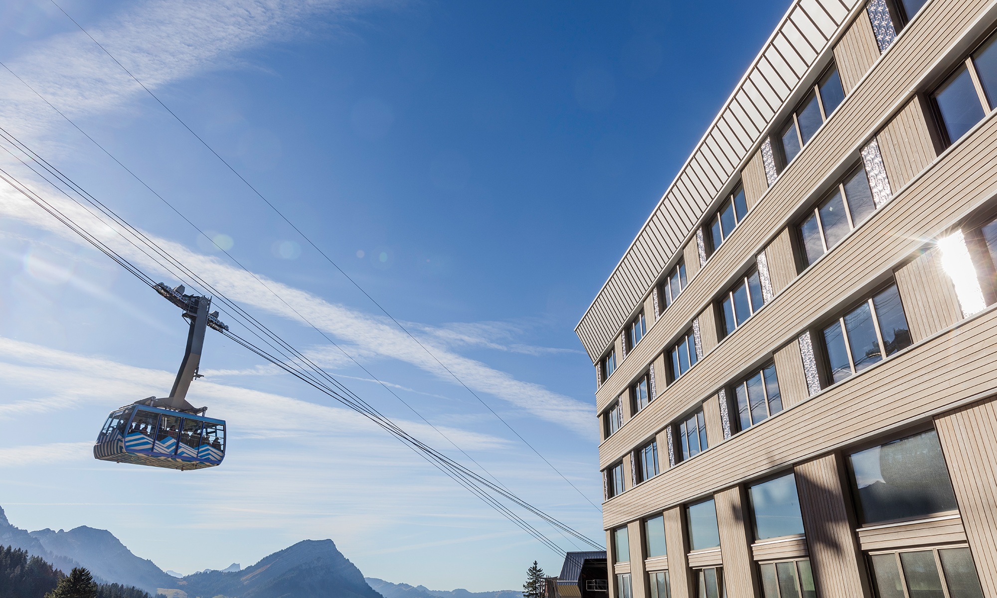 The photograph shows the facade of the Hotel Säntis and the cable car with passengers. In the background are panoramic mountain views with a radiant blue sky.