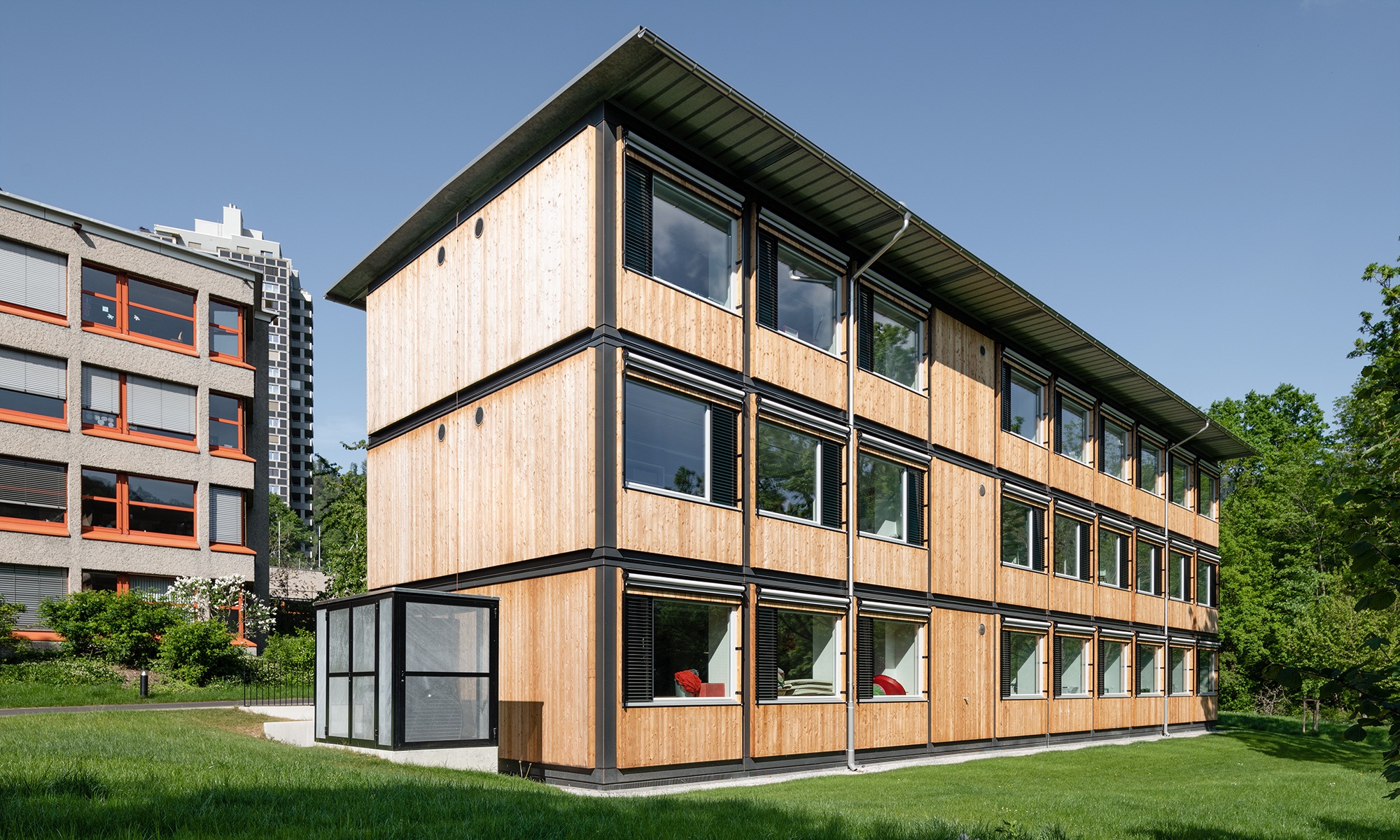 Gesamtansicht des ZM10-Schulpavillons Sihlweid in Zürich auf dem Schulareal und mit Schulhaus im Hintergrund
