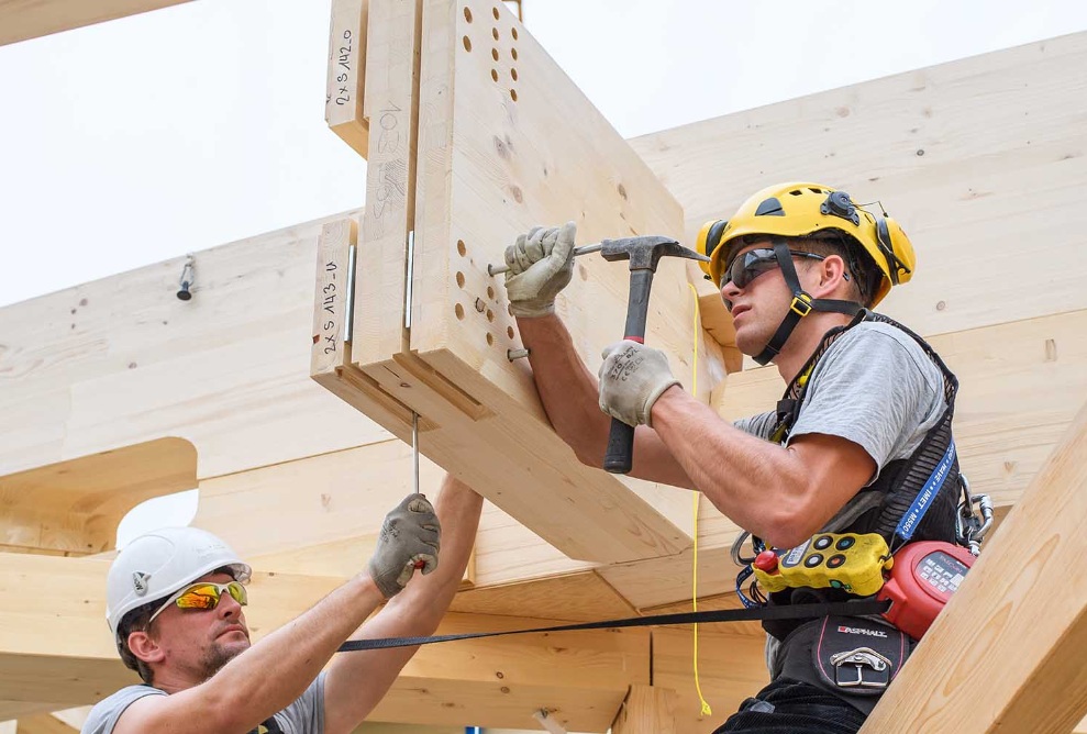 Two workers with helmets sit on a timber construction hammering in nails.