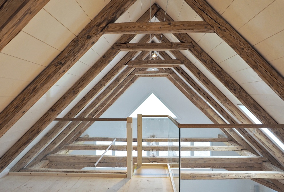 Open attic bathed in light, secured with glass and wooden guardrails. The roof slopes are clad with wide wooden beams.