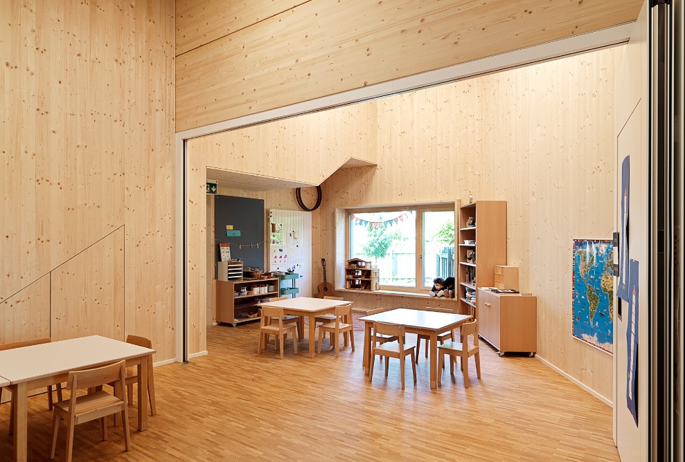 Vue intérieure de la salle de classe dans le jardin d’enfants de Wittenbach après transformation. Les murs, le plancher et le plafond de la salle sont en bois. La salle peut être divisée en deux pièces grâce à une cloison pliante. 