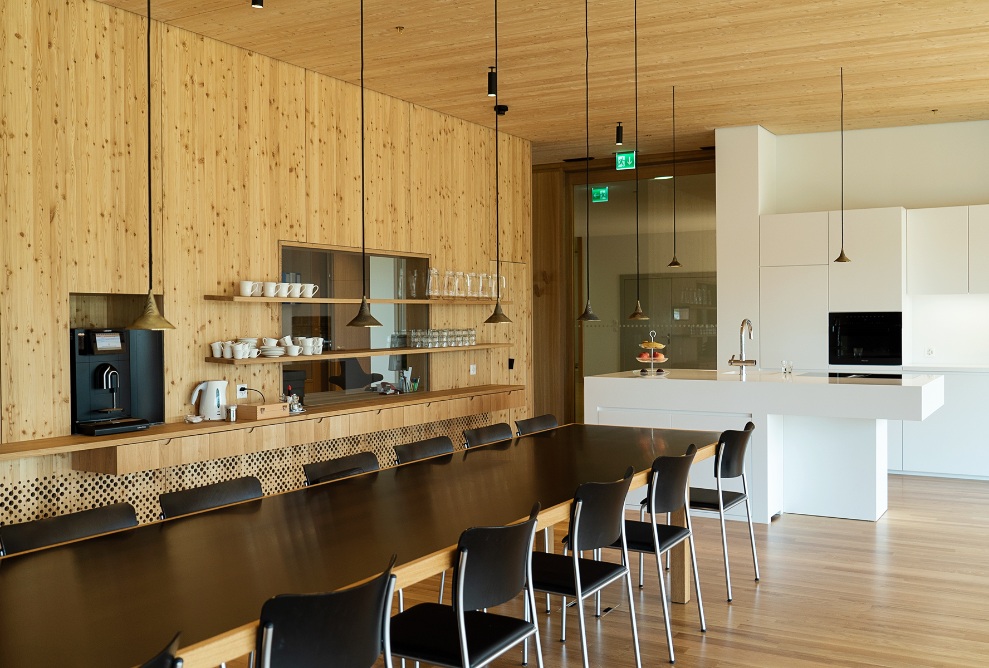 Vue intérieure de la cuisine avec de nombreux éléments en bois, un îlot, une longue table noire et des armoires blanches.
