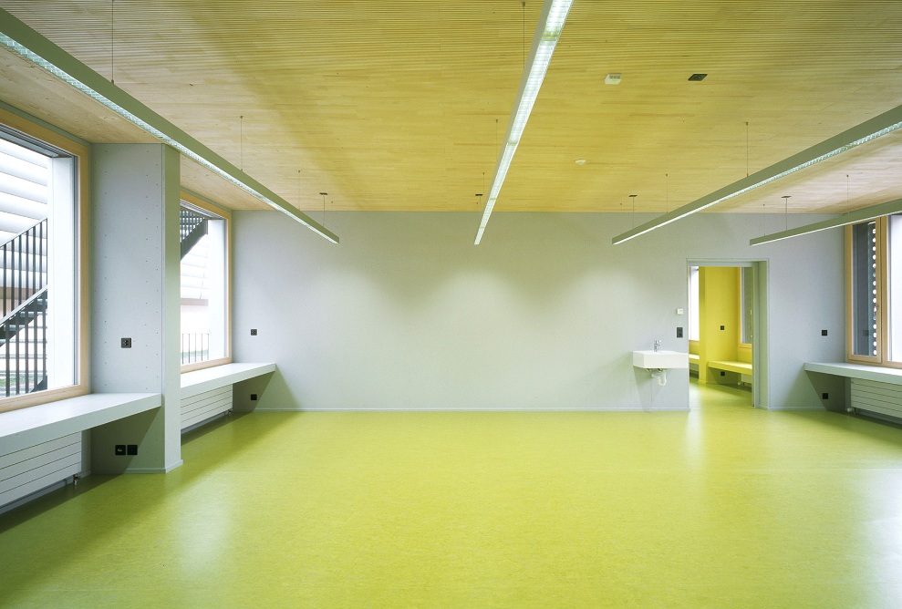 Vue sur une salle de classe vide avec des fenêtres des deux côtés, un sol jaune et un plafond en épicéa.