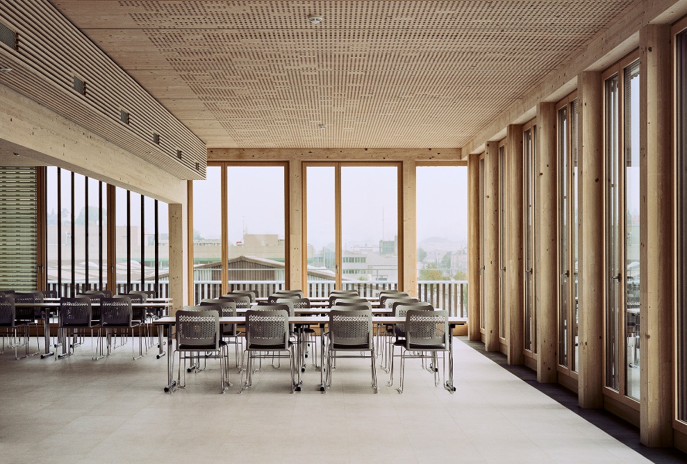 The lounge area is furnished with long tables and seating. The space is flooded with light thanks to the full-length windows while the walls and roof structure are made entirely of wood.