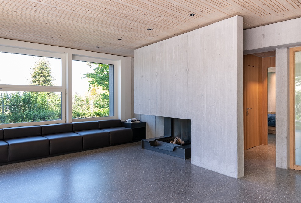 Lounge in Sutra House with large windows, concrete walls, wooden ceiling and doors plus a sofa and fireplace. 
