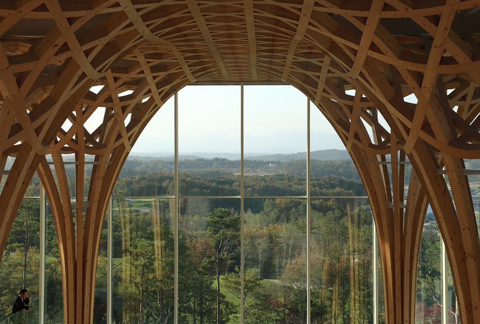 Vue depuis l’entrée à travers la façade vitrée opposée sur le terrain de golf. La structure porteuse en bois domine l’espace.
